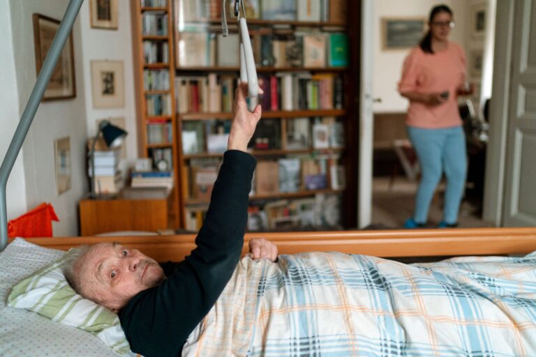 Pflegebett auf Krankenkasse – Senior im Pflegebett mit Aufrichthilfe, Pflegekraft im Hintergrund